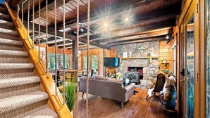 A wooden staircase with a rope handrail, a stone fireplace, a gray sofa, and a television mounted on the wall. - Riverfront Cabin with heated pool, Cedar wood Hot Tub, Dry Sauna, and Dock (Waterford)