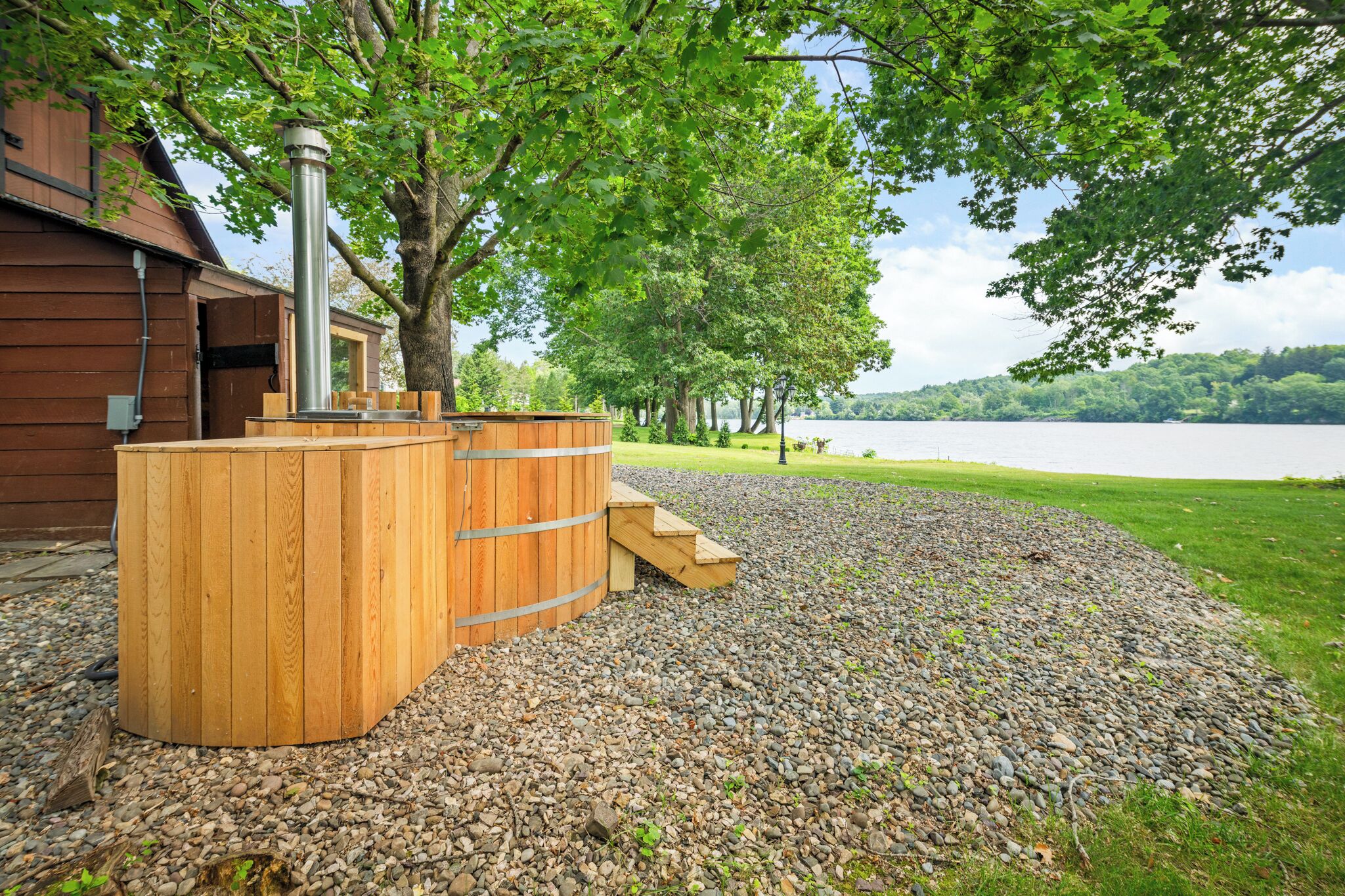 A wooden sauna structure with a metal chimney, surrounded by a gravel area and trees, near a wooden building by a lake.
