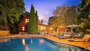 A swimming pool with lounge chairs and an umbrella, surrounded by trees and a wooden house. - Riverfront Cabin with heated pool, Cedar wood Hot Tub, Dry Sauna, and Dock (Waterford)