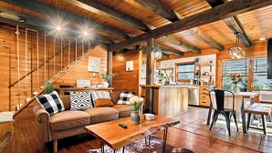 A cozy cabin interior with a wooden staircase, a living area with a sofa and coffee table, a kitchen with a bar, and a large window offering a view. - Riverfront Cabin with heated pool, Cedar wood Hot Tub, Dry Sauna, and Dock (Waterford)