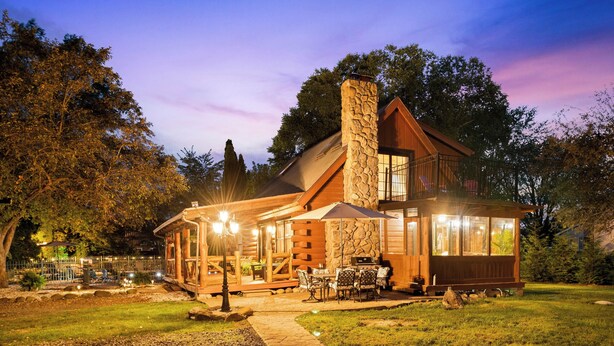 A log cabin with a stone chimney, outdoor patio, and a well-lit dining area. - Riverfront Cabin with heated pool, Cedar wood Hot Tub, Dry Sauna, and Dock (Waterford)