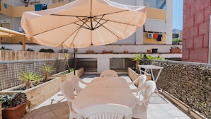 Une terrasse avec une table et des chaises, un parasol, des plantes en pot et un bâtiment en arrière-plan.