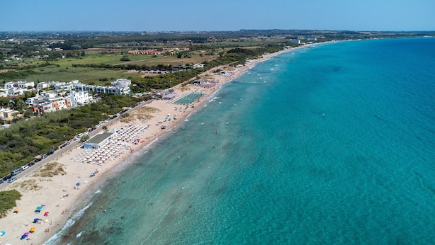 Vue aérienne d’une zone côtière avec une plage, des bâtiments et une eau d’un bleu limpide.
