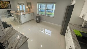 A modern kitchen with a marble countertop, white cabinets, and a large window providing natural light.