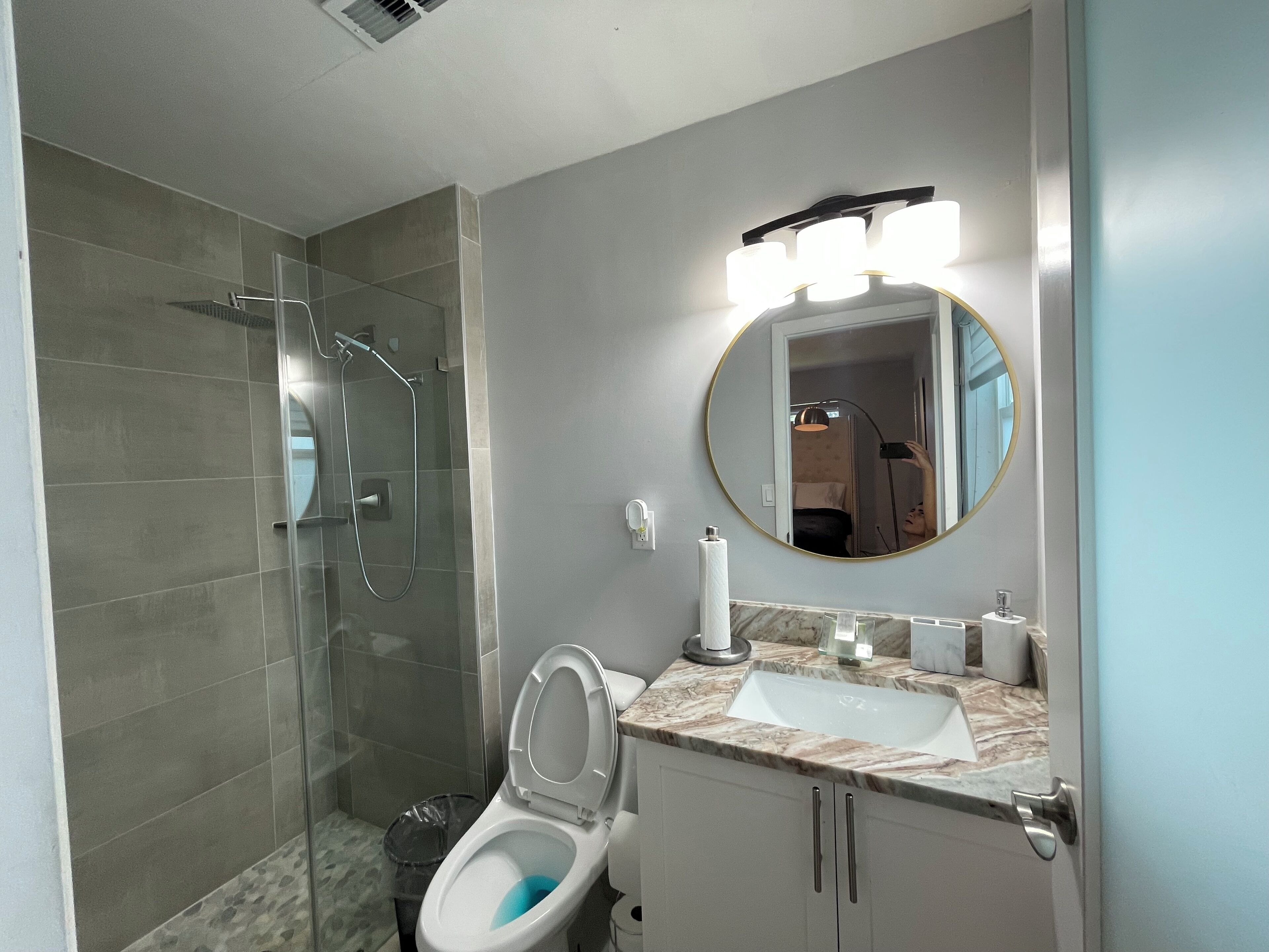 A modern bathroom with a glass-enclosed shower, a white toilet, a marble sink, and a round mirror with overhead lights.