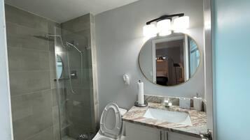 A modern bathroom with a glass-enclosed shower, a white toilet, a marble sink, and a round mirror with overhead lights.