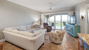 A living room with a sectional sofa, a patterned ottoman, a ceiling fan, and a view through a glass door to the outdoors. - Fall Deals + Snowbird Rates | Ocean Views + Beach Getaway (Flagler Beach)