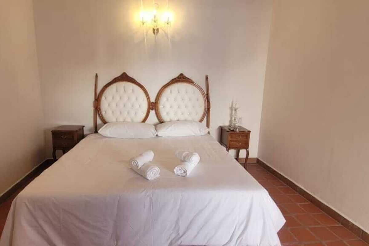 Una cama bien tendida con cabecera de madera, dos mesitas de noche y una lámpara fijada a la pared.