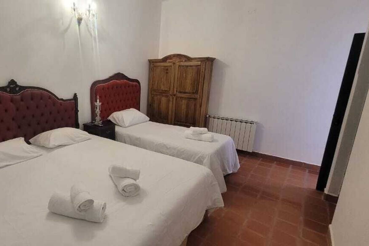Habitación con dos camas, cada una con cabecero rojo y ropa de cama blanca. Se observa un armario de madera y un radiador en la pared.