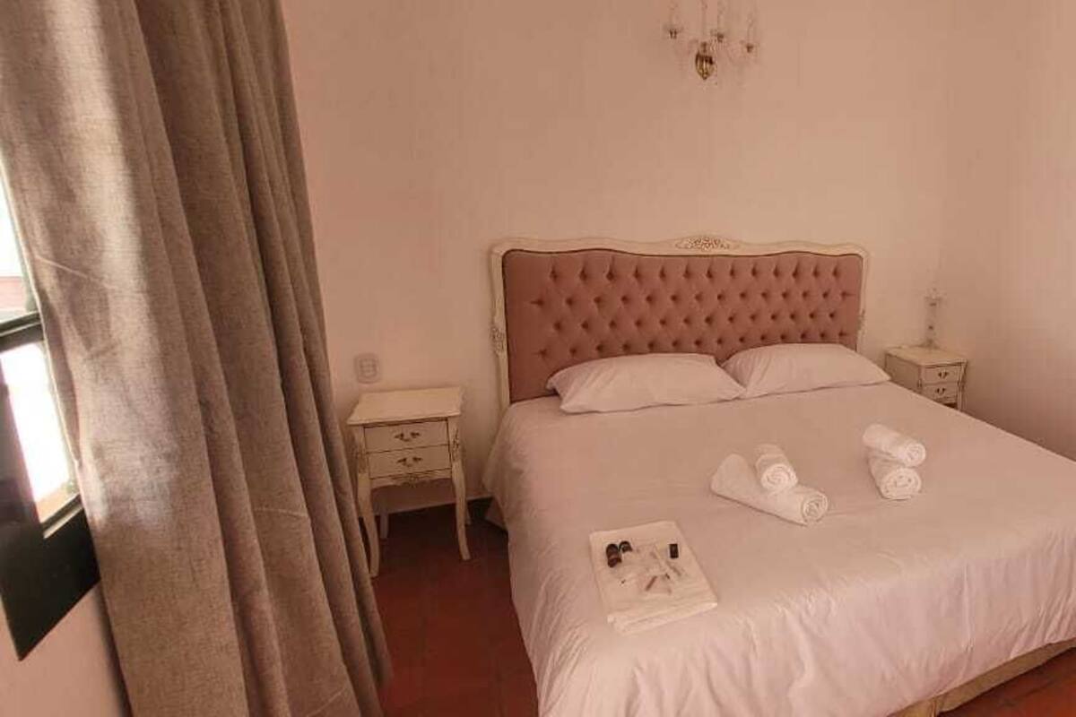 Una cama bien hecha con toallas enrolladas, una mesita de noche con lámpara y una ventana con cortinas.