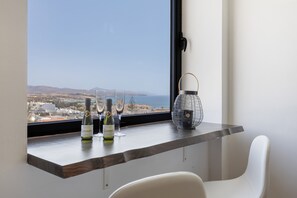 A room with a view of the sea, a table with wine bottles and glasses, and a lamp.