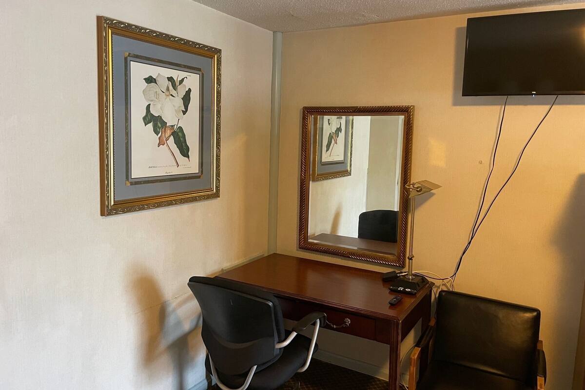 Habitación de hotel con un escritorio de madera, dos sillas de oficina negras, un espejo grande y un televisor de pantalla plana montado en la pared.