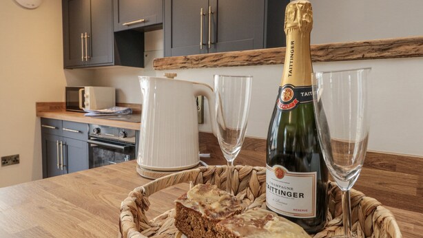 A kitchen with dark cabinets, a microwave, a white pitcher, a bottle of champagne, two glasses, and a woven basket with a slice of cake. - Betsy's Burrow (Bridlington)