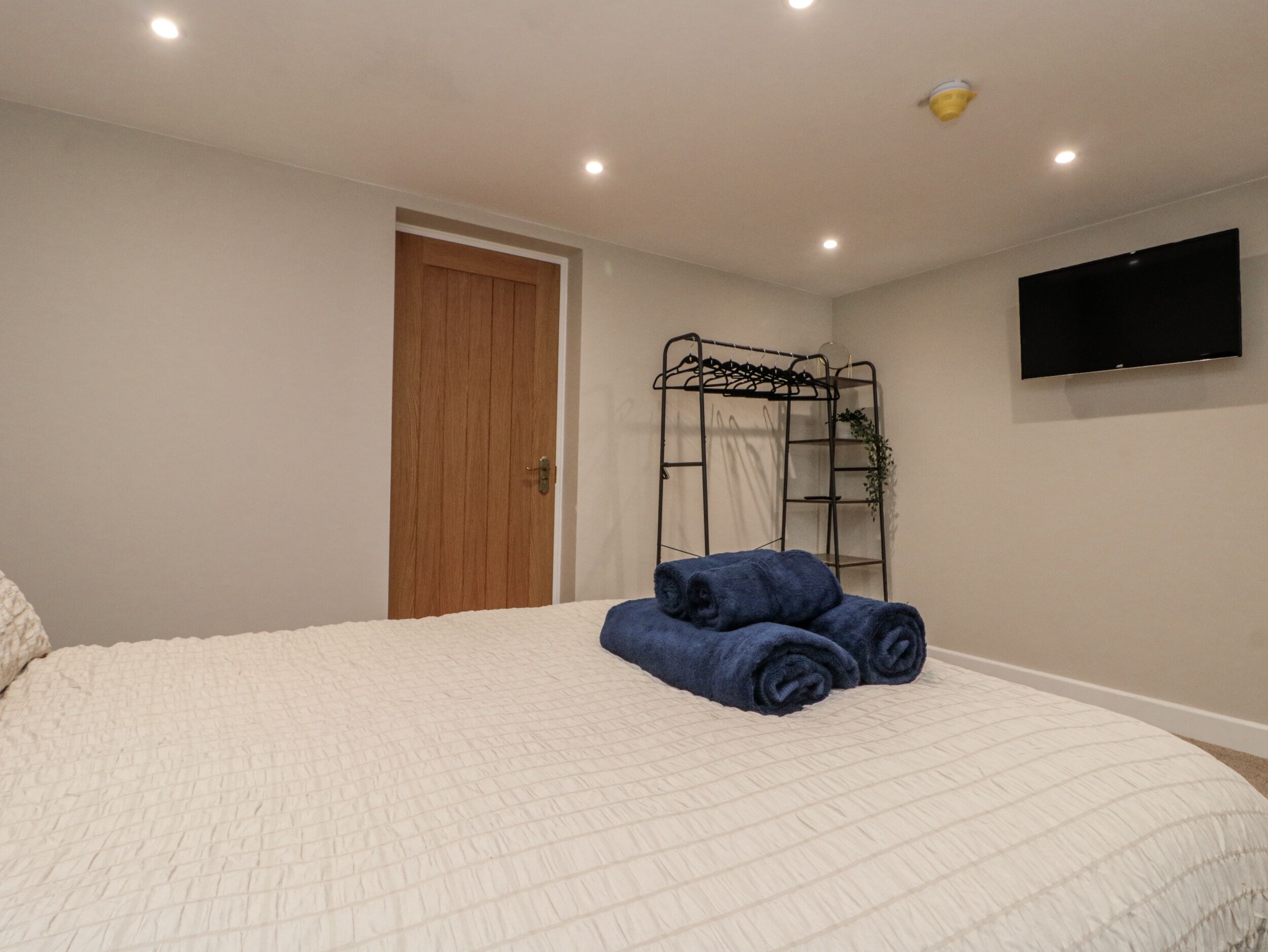 A bedroom with a bed, a wooden door, a black metal shelf, and a flat-screen TV on the wall.