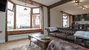 A spacious living room with a leather sofa, wooden beams, a large window, and a kitchen in the background. - Betsy's Burrow (Bridlington)
