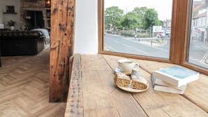 A wooden bench with a cup of coffee, a plate of cake, and books by a window overlooking a street. - Betsy's Burrow (Bridlington)