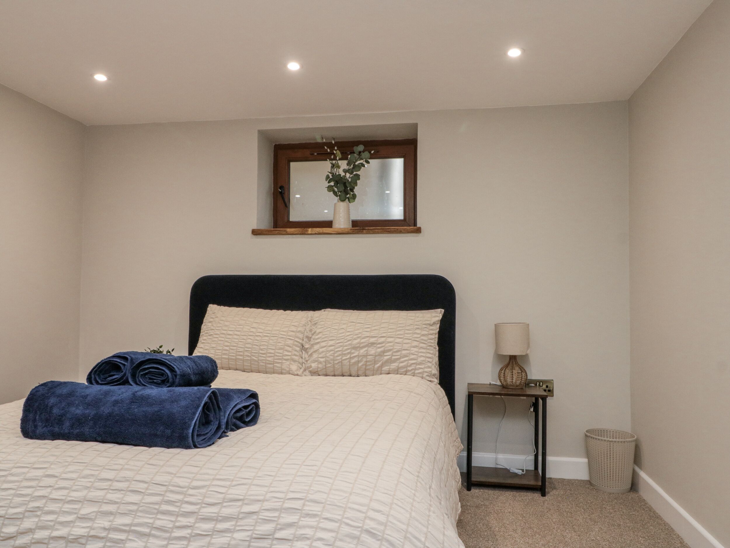 Un dormitorio con cama, mesita de noche, lámpara y un ventanal pequeño.