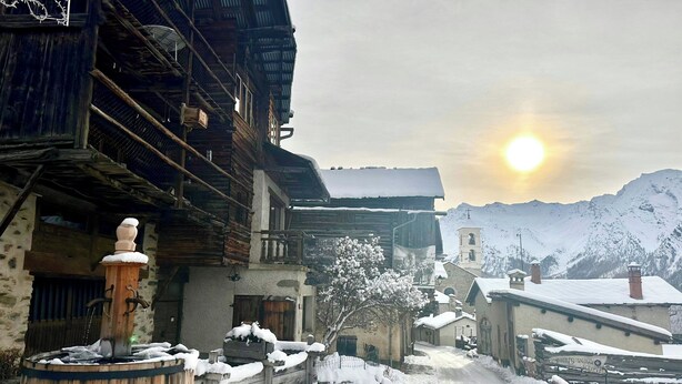 Calle nevada en un pueblo con edificios de madera, un pozo de agua de madera y una montaña al fondo.