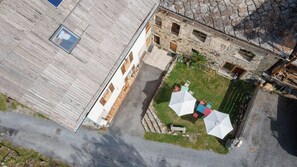 Eine Vogelperspektive zeigt einen Innenhof mit einem Steinhaus, einer Holzterrasse und Gartenmöbeln mit Sonnenschirmen.