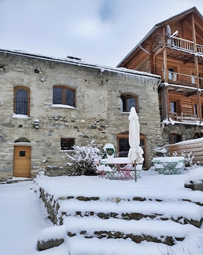 A stone building with a wooden balcony, a door, and a small garden area with a table and chairs covered in snow.
