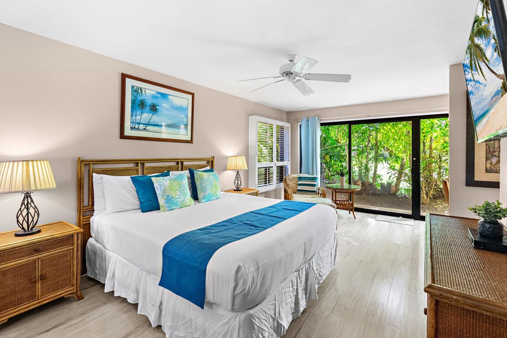 A spacious bedroom with a large bed, two bedside tables, a ceiling fan, a window with a view of greenery, and a painting on the wall.