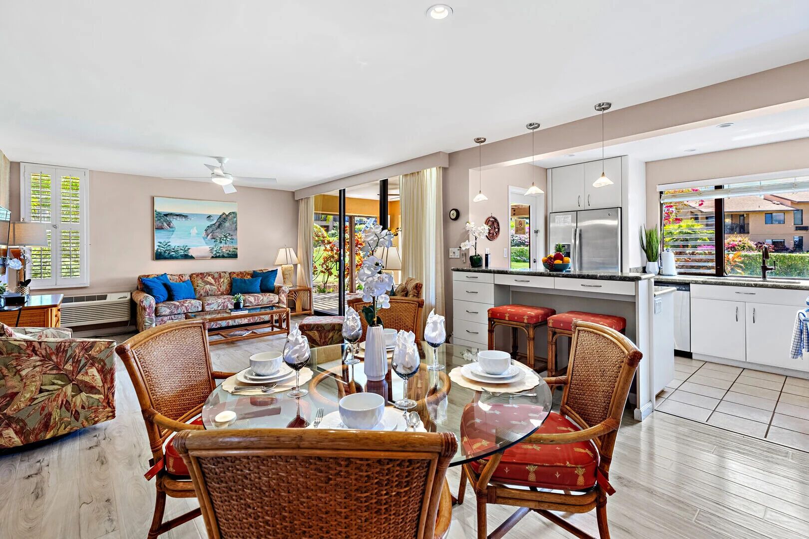 A spacious living area with a dining table set for four, surrounded by wicker chairs. A sofa with blue cushions is positioned against the wall, and a painting is visible above it. The kitchen area is equipped with modern appliances and a large window offering a view of the outdoors.