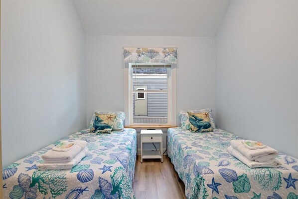 Un alojamiento pequeño de una sola habitación con dos camas individuales, cada una con ropa de cama floral a juego. Se ve una ventana con cortina y una pequeña mesa entre las camas.