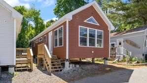 A single-story mobile home with a front porch and a small deck, surrounded by trees and other mobile homes.