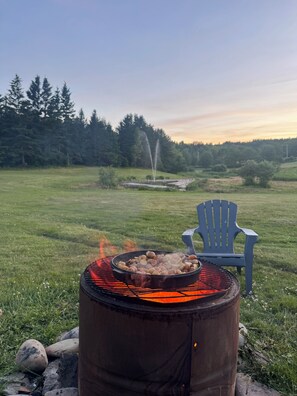 A portable grill with food, a blue chair, and a natural landscape with trees and a fountain. - 2 king BD Full Kitchen Large Outdoor Space | Unit 14 @ Great Cabot Lodge (Baddeck)