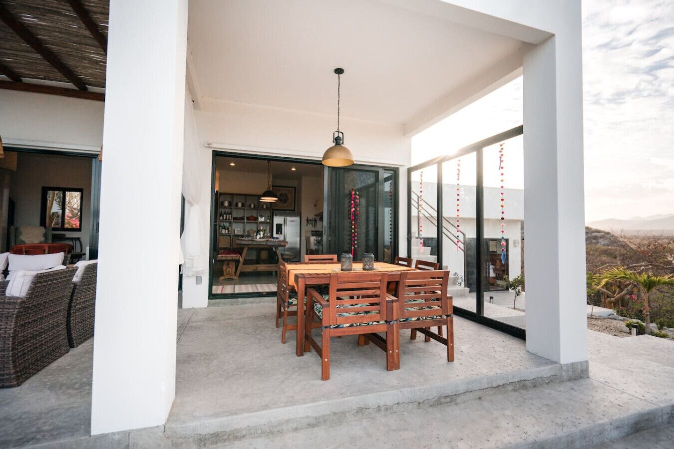 Un espace de restauration extérieur abrité, avec une table et des chaises en bois, un luminaire suspendu et une vue sur l’intérieur à travers des portes vitrées.