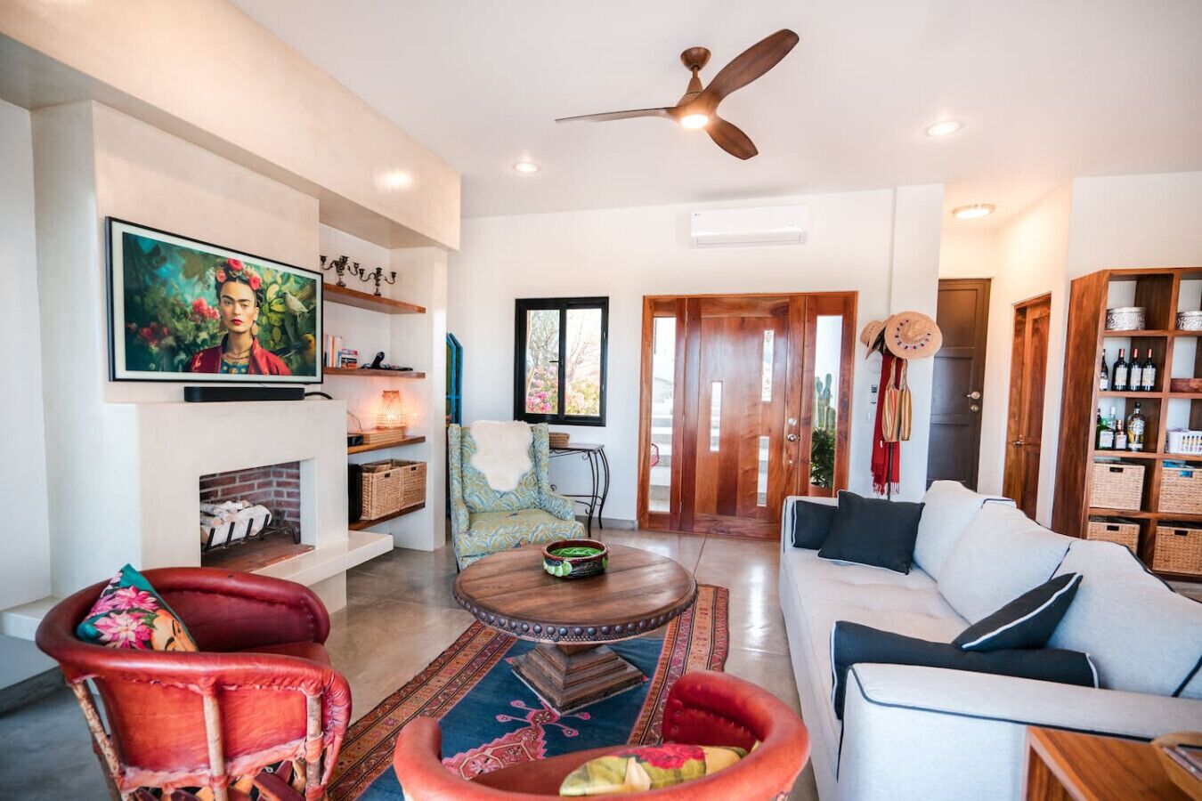 A spacious living room with a sofa, armchairs, a coffee table, and a fireplace. There is a ceiling fan, a television mounted on the wall, and a bookshelf.