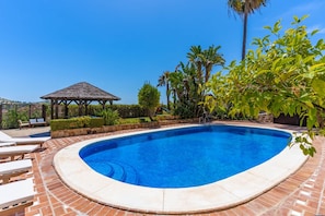 A swimming pool with a curved edge, surrounded by a brick patio, a thatched-roof gazebo, and lush greenery.