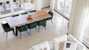 A modern dining area with a wooden table and green chairs, a white sofa with decorative pillows, and large windows offering a view of outdoor furniture. - Villa Oriente - Family Villa with pool in Marbella East (Marbella)