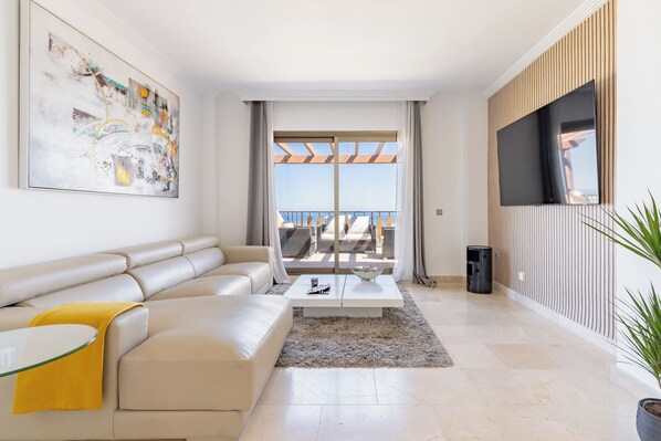 Un salon moderne avec un canapé beige, une table basse en verre et un grand tableau accroché au mur. Il y a un balcon avec des meubles d’extérieur et un téléviseur fixé au mur.