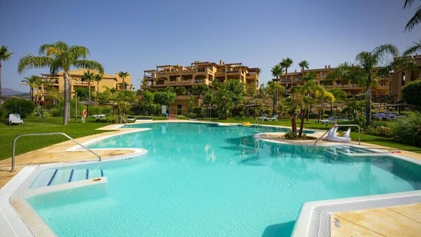 A swimming pool with a clear blue water, surrounded by palm trees and a building with multiple balconies in the background. - Penthouse 2BR with Sea Views & Heated Pool (Benahavís)