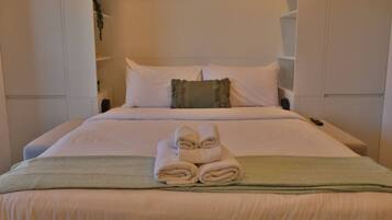 Una cama bien hecha con una toalla doblada al frente, ropa de cama blanca y un plaid verde.