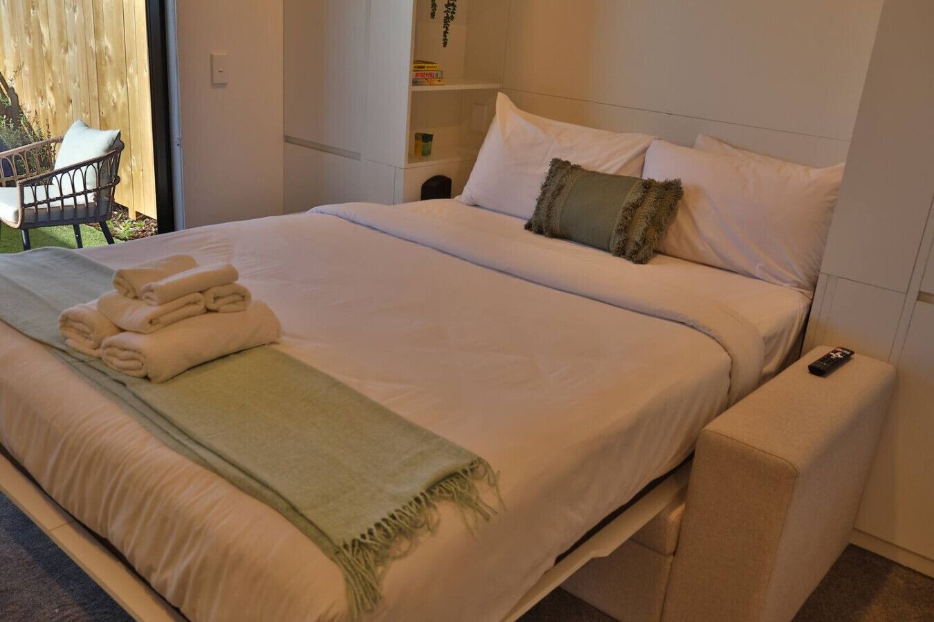Una cama bien hecha con toallas dobladas y una manta verde, una mesita de noche con un control remoto, y una silla fuera de la habitación.