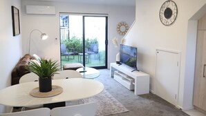 Ett modernt vardagsrum med en matplats, en tv på ett vitt tv-bord och ett stort fönster med utsikt över en trädgård.
