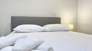 A neatly made bed with white linens and pillows, a grey headboard, and a lamp on the nightstand.