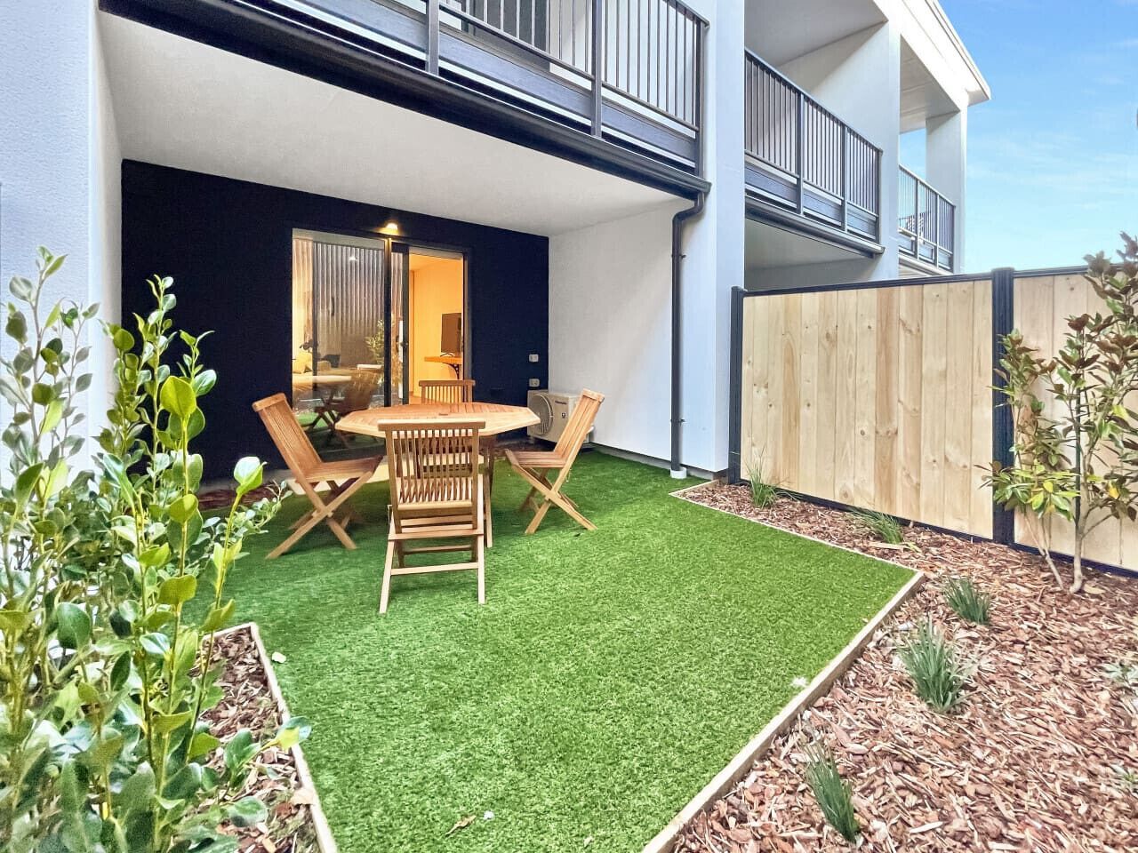 A modern outdoor patio with a round wooden table and chairs, surrounded by artificial green grass and small plants.