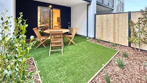 Une terrasse extérieure moderne, dotée d’une table ronde en bois et de chaises, entourée de gazon synthétique et de petites plantes.