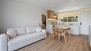 Un salon moderne avec un canapé et une table à manger, une cuisine avec des meubles blancs et une fenêtre donnant sur un paysage verdoyant.
