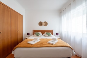Una habitación de hotel con cama, armario de madera, dos mesitas de noche y una ventana con cortinas de gasa.