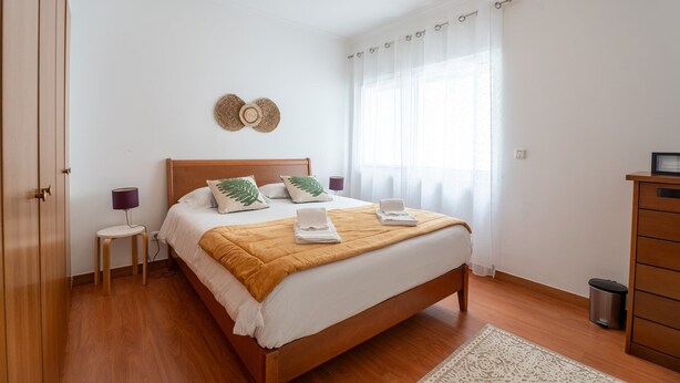A bedroom with a wooden bed, a bedside table, a lamp, a trash bin, and a wardrobe.
