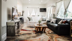 Un salon moderne avec un canapé, une table basse et un téléviseur. Il y a également un coin cuisine avec un bar et des tabourets. La pièce est dotée de grandes fenêtres avec des rideaux.
