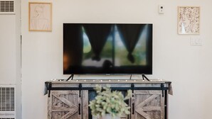 Un salon avec un téléviseur à écran plat fixé à un meuble en bois, une table basse en verre sur laquelle se trouve un vase, et deux œuvres d’art encadrées accrochées au mur.