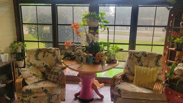 A room with two floral-patterned chairs, a round wooden table, and various potted plants.