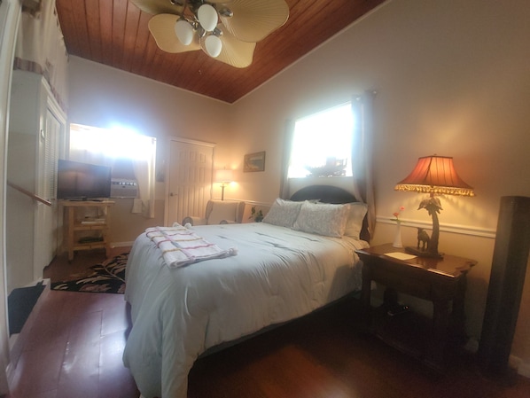 Un dormitorio con cama, lámparas de noche, ventilador de techo, ventana y televisor.