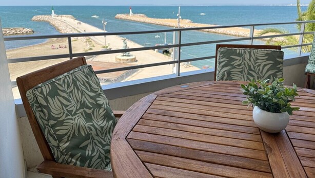 A wooden table with a potted plant, two chairs with patterned cushions, and a view of the sea and a rocky shoreline with lighthouses.