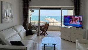 A living room with a sofa, a glass coffee table, a TV, and a view of the sea through large windows.